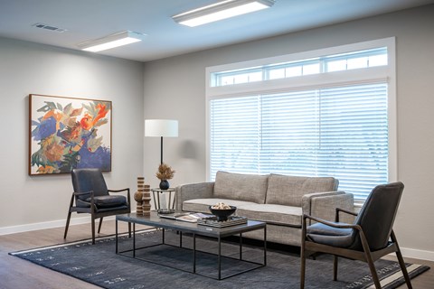 A living room with a grey couch, a black chair, a glass table, and a painting on the wall.