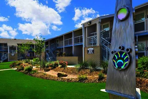 A large wooden pole with a colorful design stands in front of a building with a green lawn and trees.