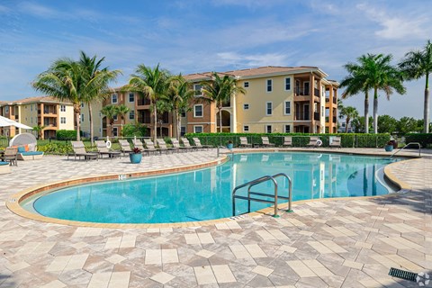 A swimming pool surrounded by a brick patio and palm trees in front of apartment buildings.