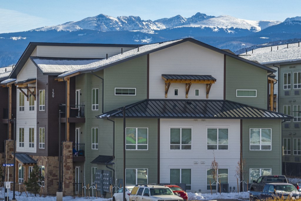 Apartment complex with cars parked in front and snowy mountains in the background.