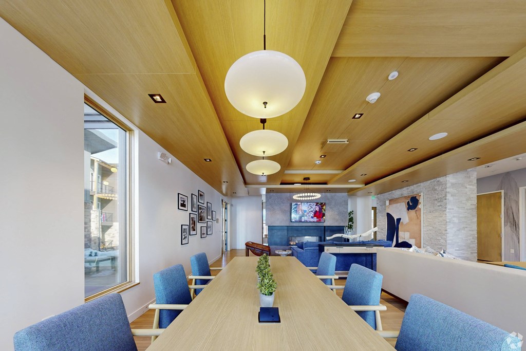 A long wooden table with blue chairs is in the middle of a room with a white wall and a TV.