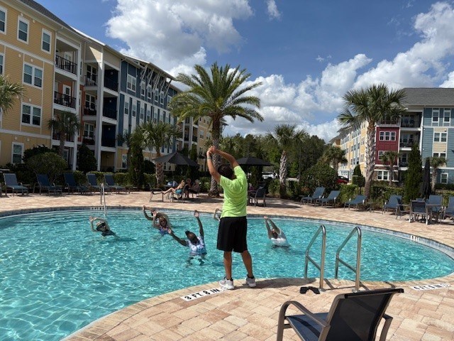 A man in a green shirt is standing by a pool with people swimming.