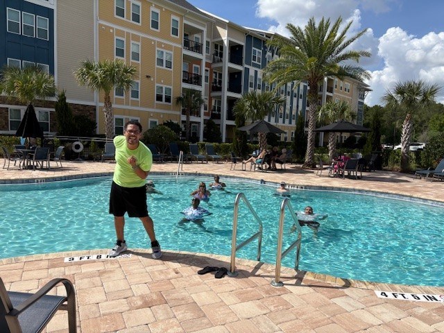 A man in a yellow shirt is standing by a pool with people swimming.