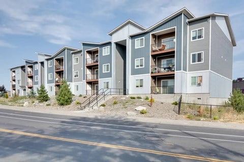 A row of modern apartment buildings with balconies and pine trees.