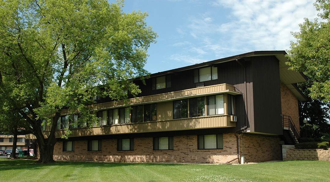 A large brown building with a balcony on the second floor is surrounded by trees.