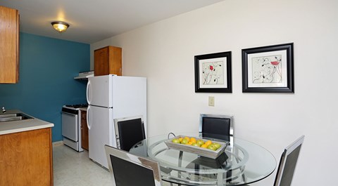 A kitchen with a white refrigerator and a glass table with two chairs and two framed pictures on the wall.