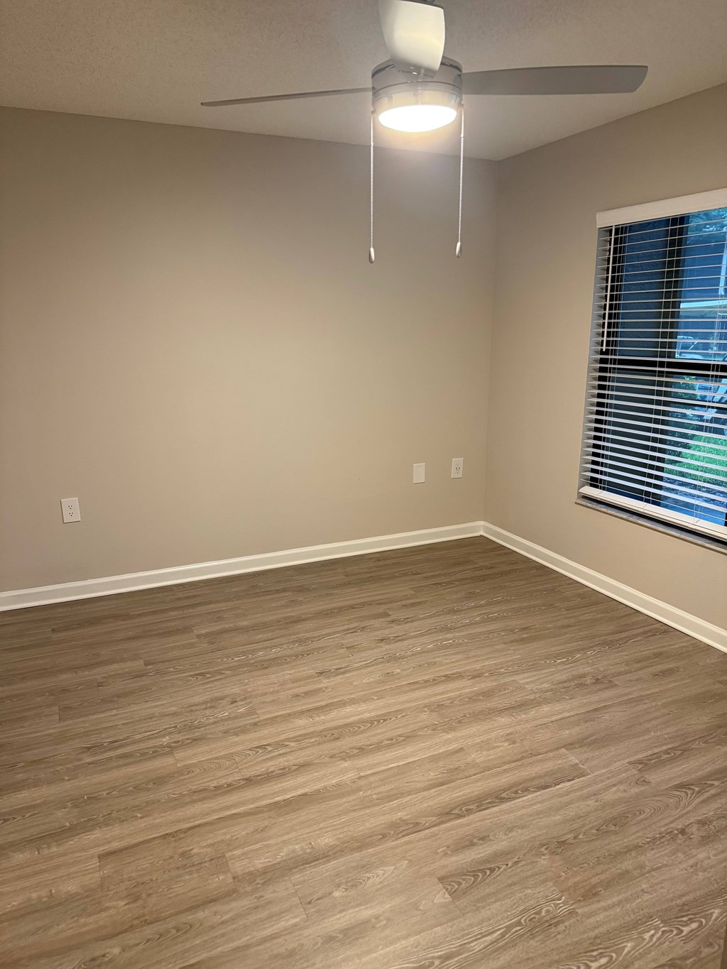 A room with a ceiling fan and light, wooden flooring, and a window with blinds.