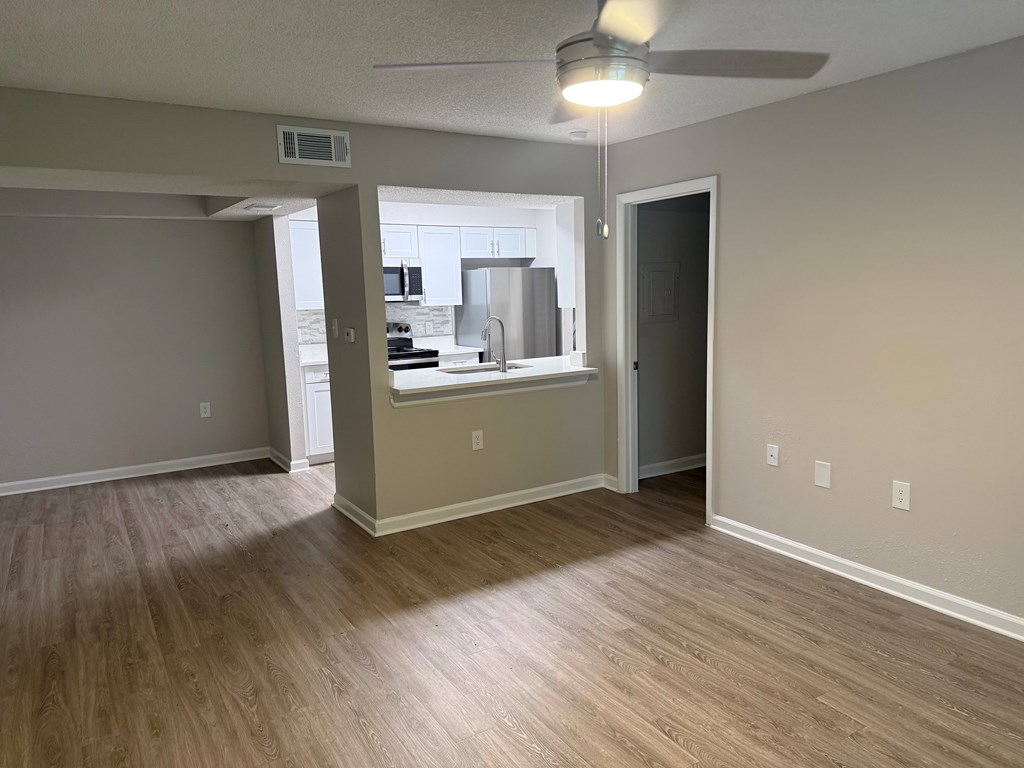 A room with a ceiling fan and light fixture, and a kitchen area with a stove and refrigerator in the background.