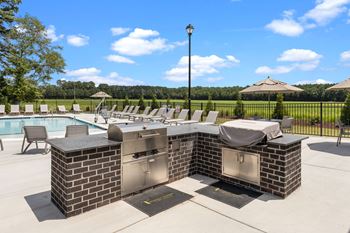 A brick barbecue grill with a table and chairs is in the foreground with a pool and trees in the background at Regency Townes in Greenville, NC