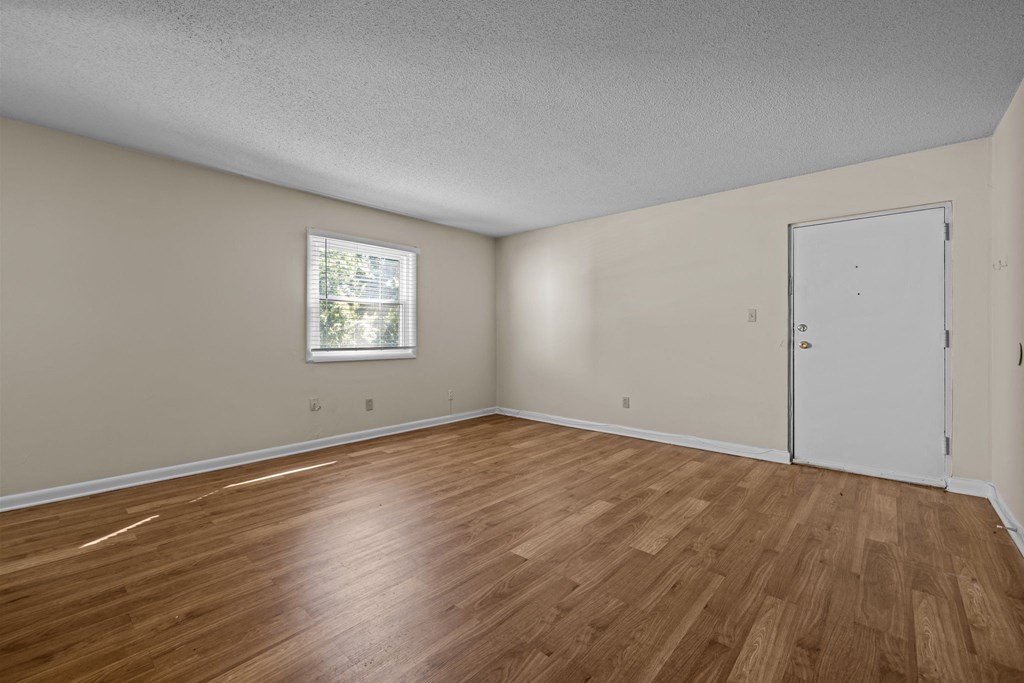 the spacious living room of an empty home with wood flooring and a window
