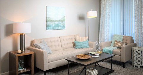 A living room with a beige couch, a wooden side table with a lamp, and a coffee table with a bowl on it.