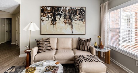 A living room with a beige couch, a painting on the wall, and a small table with a lamp.