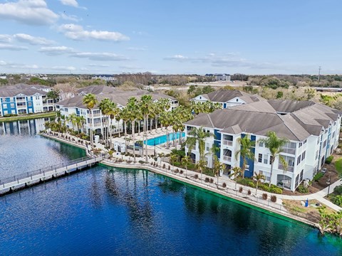 A large white building with a pool is surrounded by palm trees and a body of water.