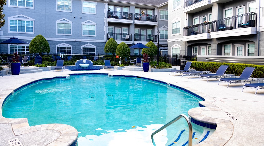 Apartments in Tanglewood, TX for Rent - Crosley Tanglewood - Pool Area with Blue Poolside Lounge Seating, Umbrellas, Tables, and Decorative Fountain