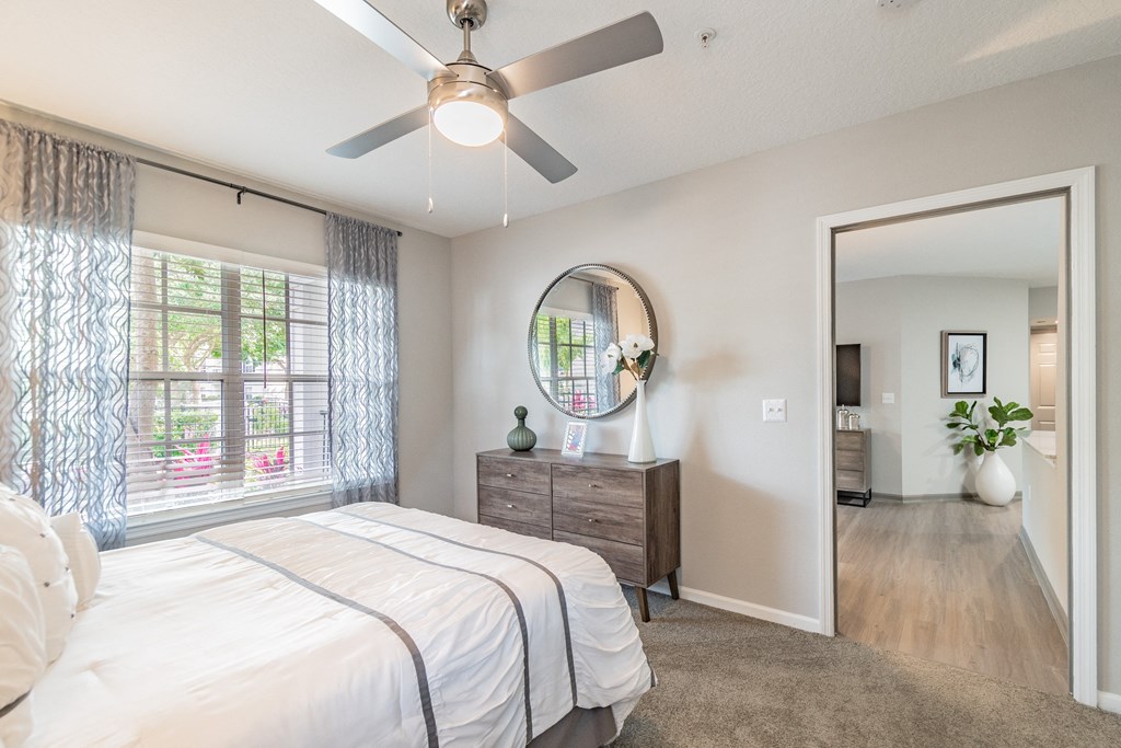 One BR Apartments in Orlando FL- Infinity off Baldwin Park - Bedroom with Carpeted Floor, Bed, Wooden Dresser, Oval Mirror, Large window with Curtains, and Ceiling Fan