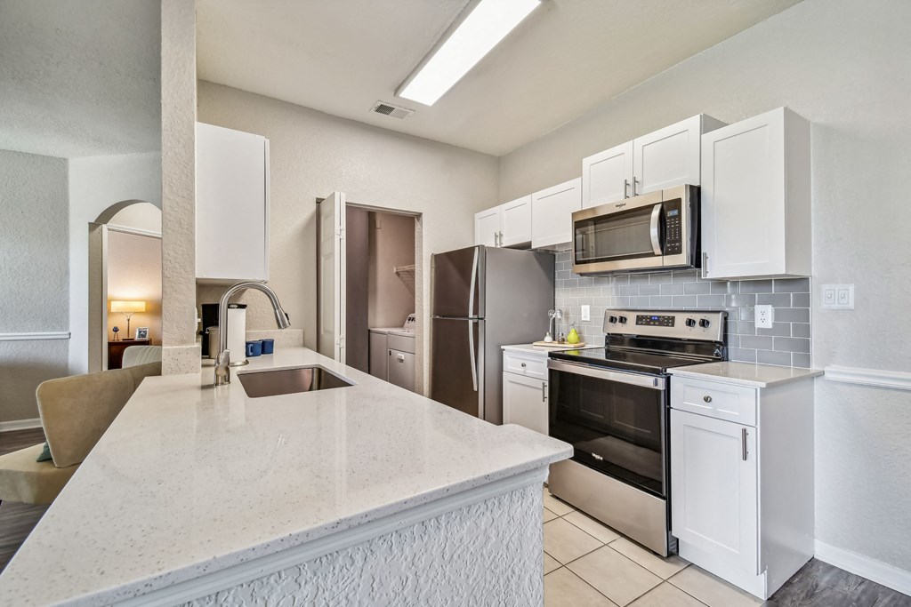 Four BR Apartments in Brandon FL - 2211 Grand Isle - Modern Aisle-Style Kitchen with Stainless Steel Appliances, White Cabinetry, Grey Tile Backsplash, Overhead White Lighting, with View of Washer/Dryer Facilities