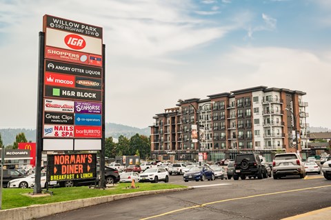 A large sign in front of a building advertises various businesses such as IGA, Shoppers, Angry Otter Liquor, Macco, H&R Block, Great Clips, Nails & Spa, and Norman