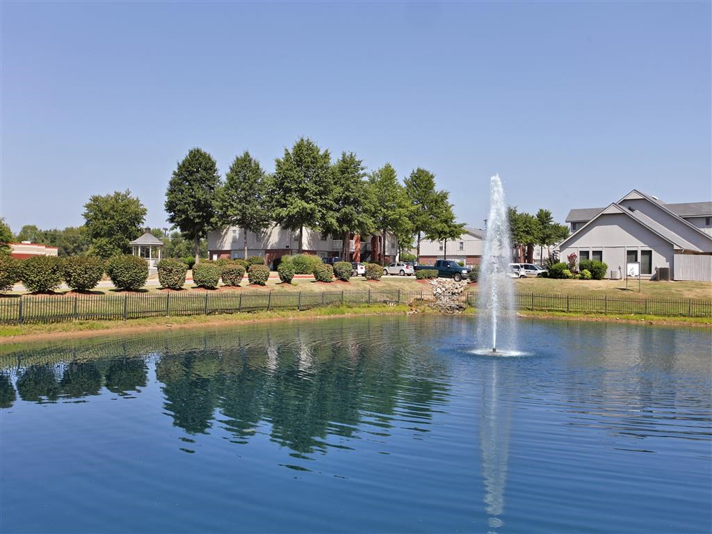 A fountain in the middle of a lake with houses in the background.