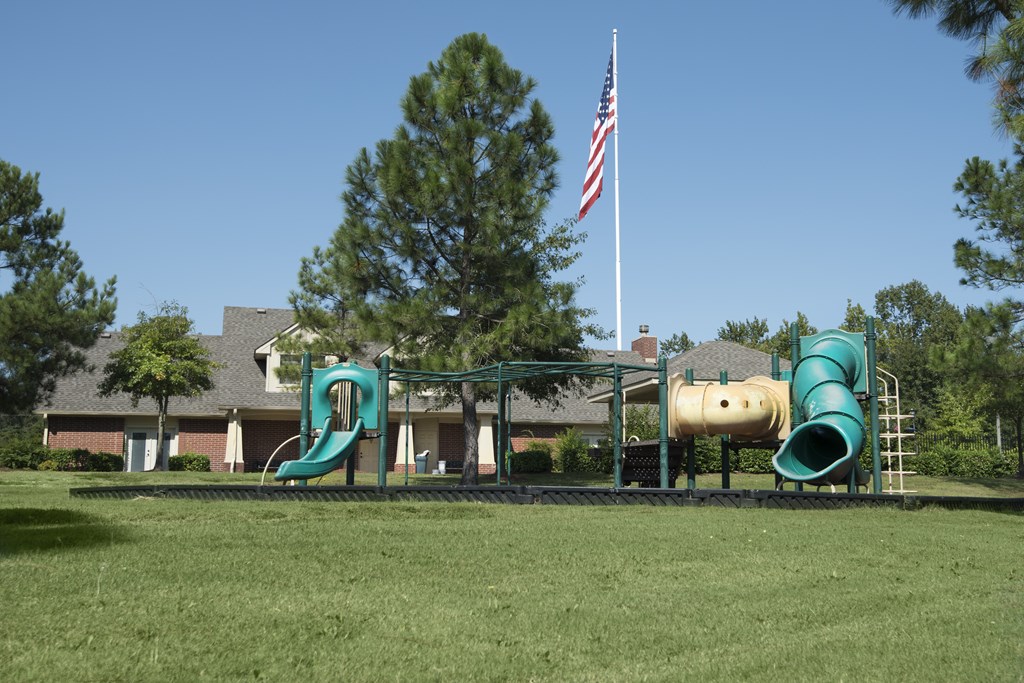 A playground with a green slide and a yellow slide in front of a house with an American flag.