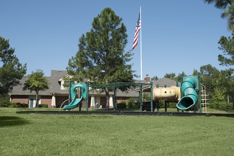 A playground with a green slide and a yellow slide in front of a house with an American flag.