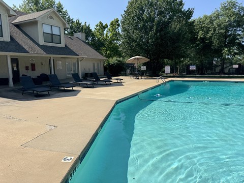 A swimming pool in front of a house with a patio and lounge chairs.