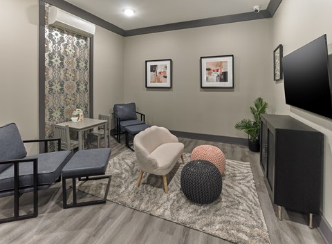 A living room with a grey carpet, a black sofa, a grey armchair, a black chair, a black ottoman, a black speaker, a black table, a black and white patterned curtain, a small table with