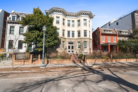 A street view of a large white building with a red building in the background.