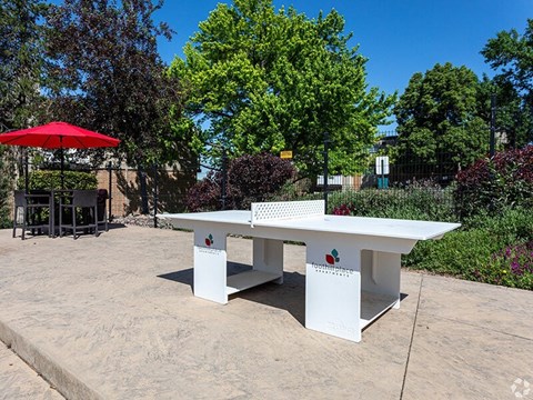 A white table with a red umbrella is in the foreground of a sunny outdoor area.