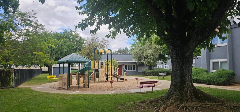 A playground with a green canopy and yellow slide is surrounded by a fence and a large tree.