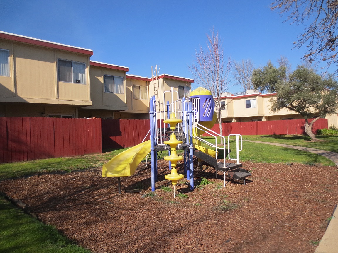 American River Gardens playground on bark with grass & concrete surrounding, in front of apartment buildings with red fencing