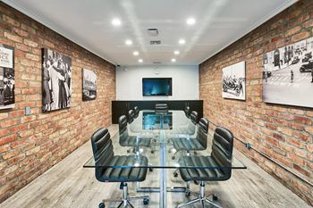 A conference room with a glass table and chairs, a television, and framed pictures on the brick walls.