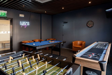 A modern game room featuring a foosball table, pool table, and shuffleboard table, with a dartboard on the wall, leather chairs, and a neon "Let's Party" sign.