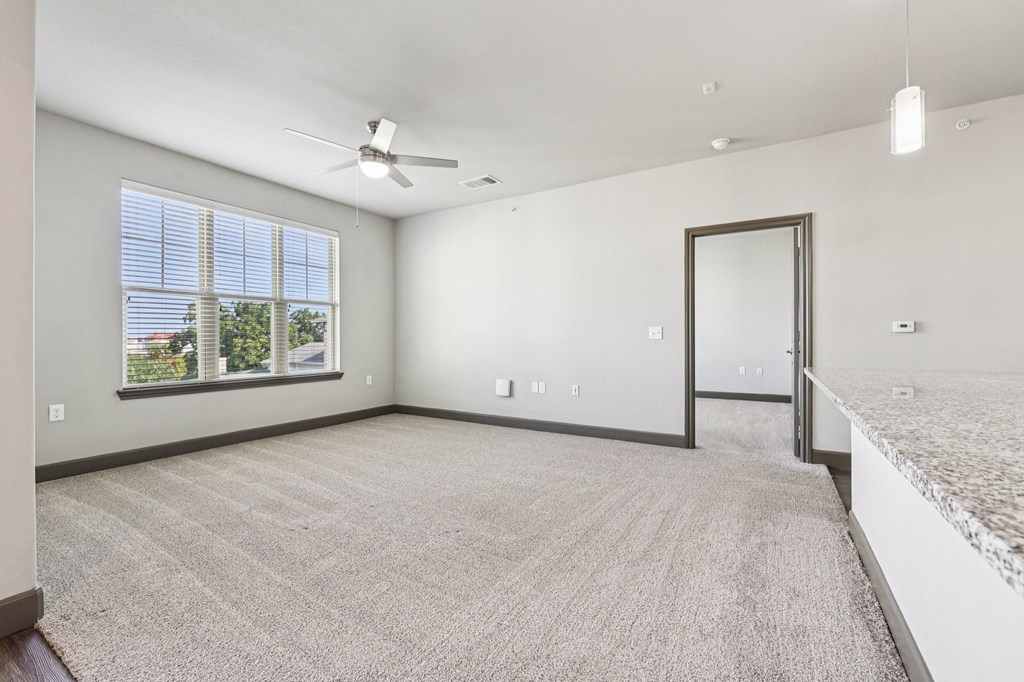 A spacious room with a carpeted floor, a ceiling fan, and a large window with blinds.