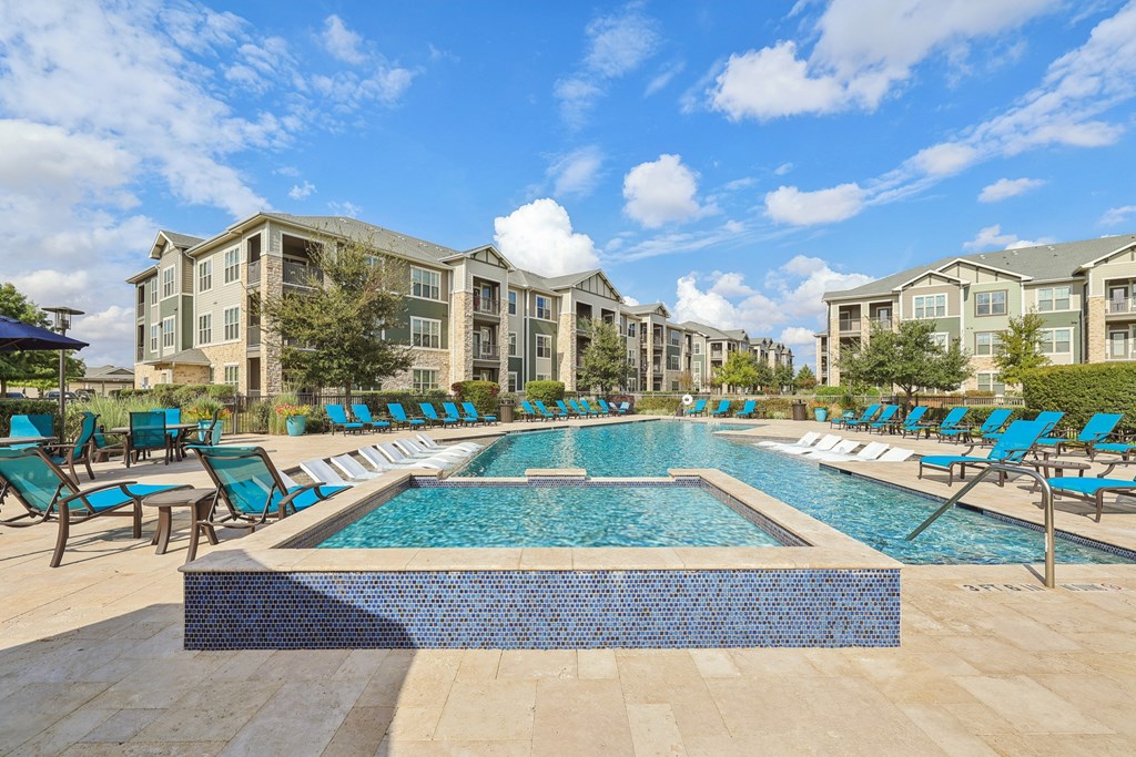 A large swimming pool surrounded by lounge chairs in front of apartment buildings.