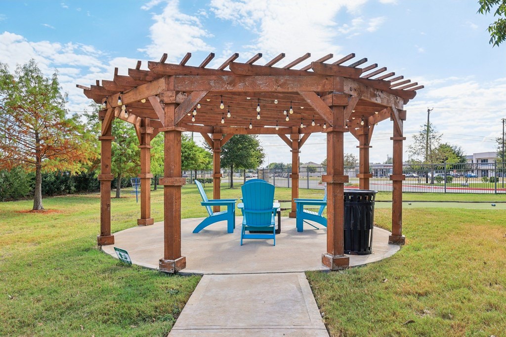 A wooden pergola with blue chairs and a trash can is in a grassy area.