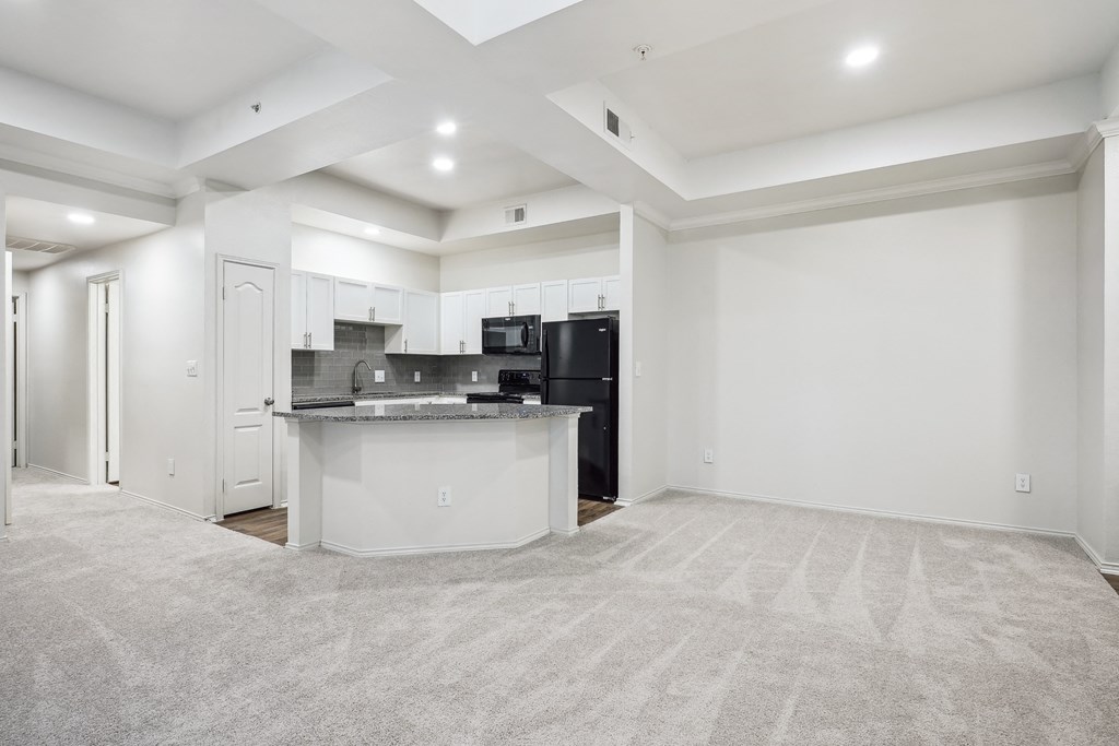 an empty living room with a kitchen with a black refrigerator at La Ventura Apartments, Texas, 75093