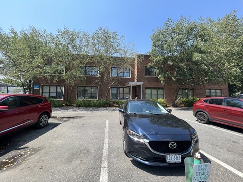 A black Mazda car is parked in a parking lot with a red car and a building in the background.