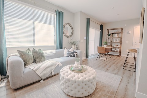 An open concept living and dining space with a white couch and a round ottoman, bar stools, dining table and bookshelf.