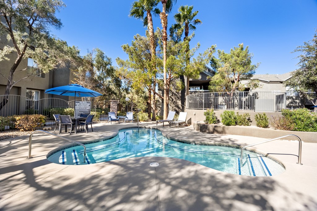 A pool surrounded by trees and chairs under a blue umbrella. at The Equestrian by Picerne Apartments, Henderson 89052