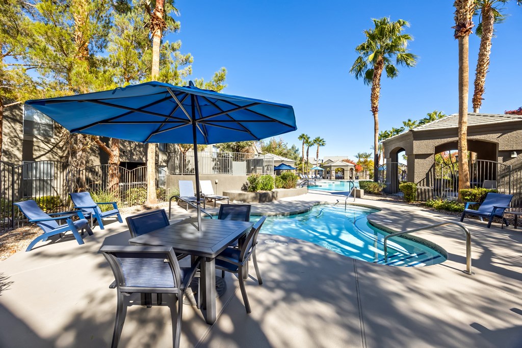 A pool area with a table and chairs and a blue umbrella. at The Equestrian by Picerne Apartments, Henderson, NV, 89052