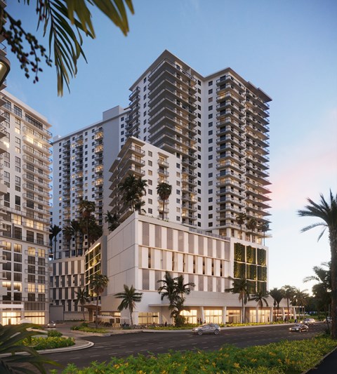 A modern building with a white facade and balconies is surrounded by palm trees and other greenery.