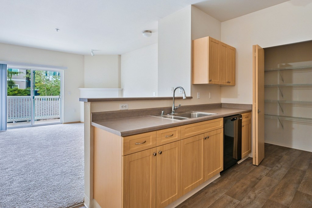 Soho kitchen with wooden cabinets, a pantry closet, and the living room in the background at Abbey Rowe Apartments in Olympia, WA