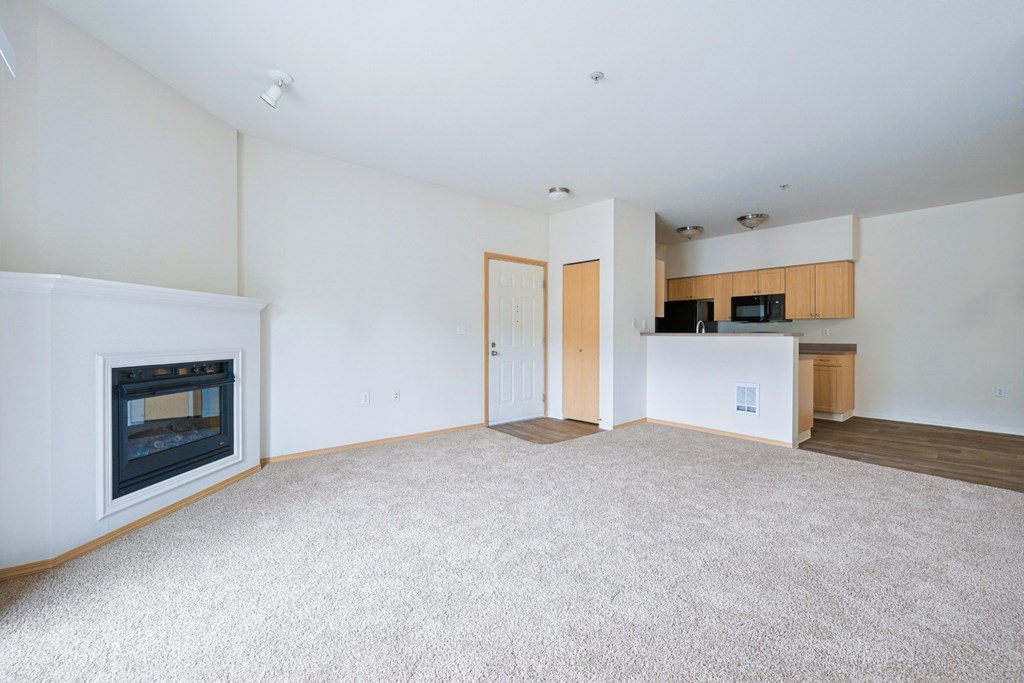 Spacious Soho living room with a fireplace, carpeting, and a kitchen in the background at Abbey Rowe Apartments in Olympia, WA
