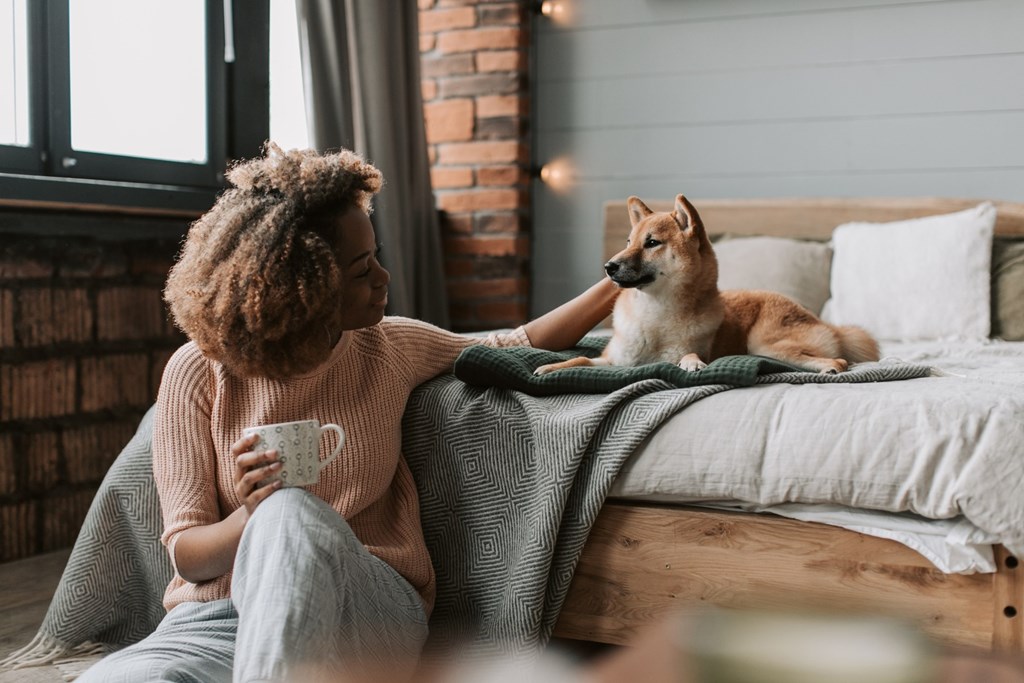 A woman with a cup in her hand is sitting on a bed with a dog beside her.
