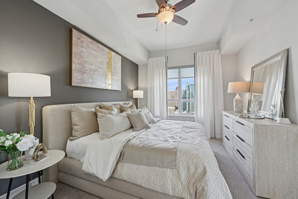 Model bedroom with a large bed, a ceiling fan, and a view of the city at Dominion Post Oak apartments in Houston, TX