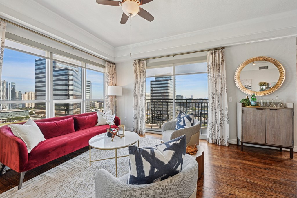 Model living room with a red couch, a white coffee table, and a large window with a view of the city at Dominion Post Oak apartments in Houston, TX