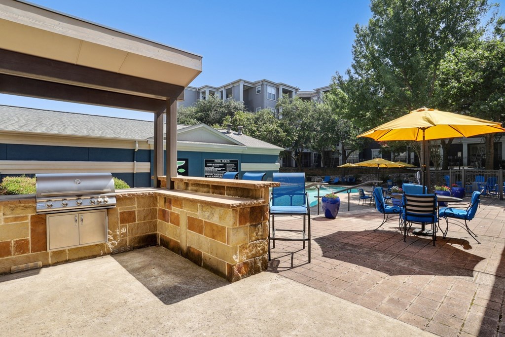 A grilling area is on a patio with blue chairs and yellow umbrellas with the pool in the background at Encore at Buckingham Apartments in Richardson, TX
