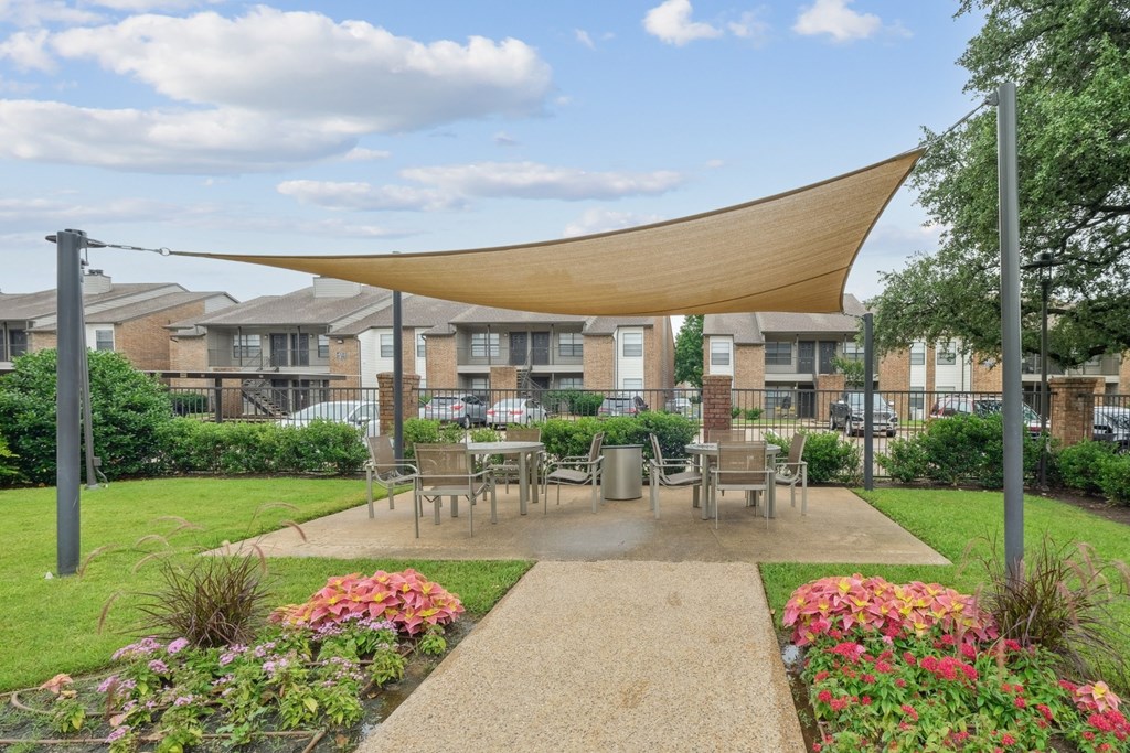 A shaded, outdoor dining area with lush grass and flowers surrounding it at Estelle Creek North Apartments in Irving, TX