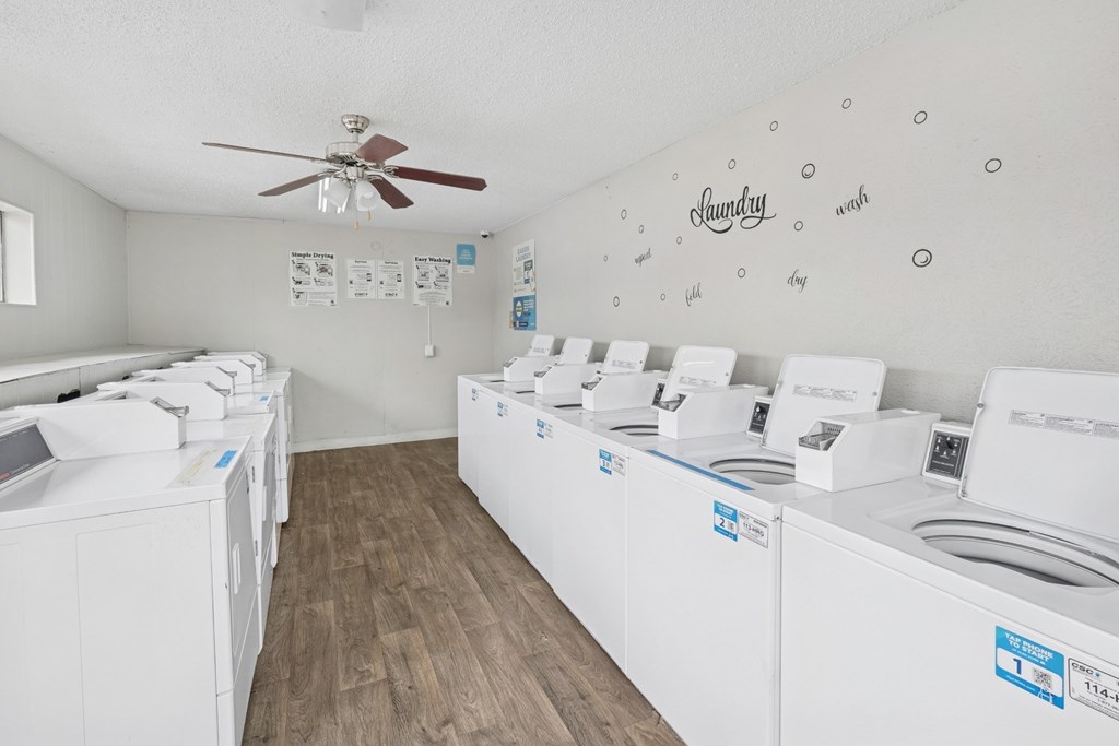 A room with a white ceiling and floor with a wall of washing machines on the right and a fan on the ceiling.