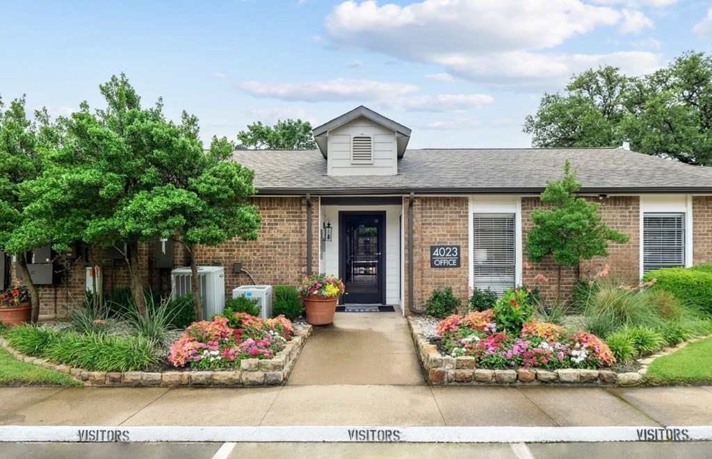 A leasing office with lush trees and blooming garden beds in front at Estelle Creek North Apartments in Irving, TX